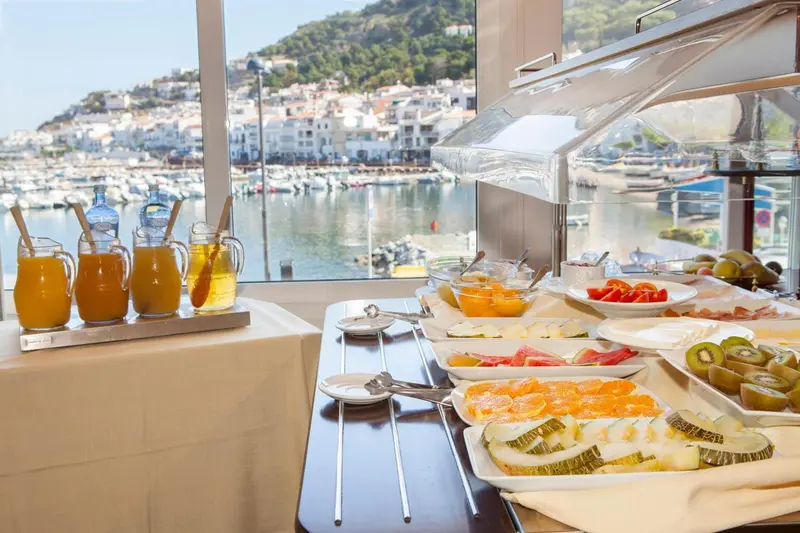 Un buffet propose des jus de fruits, des pâtisseries et des fruits frais devant une vue sur un port de plaisance.