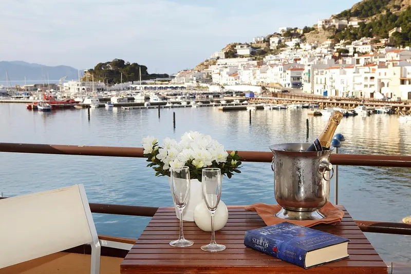 Une table en bois avec des verres à champagne, une bouteille et un livre, sur une terrasse avec vue sur un port de plaisance.