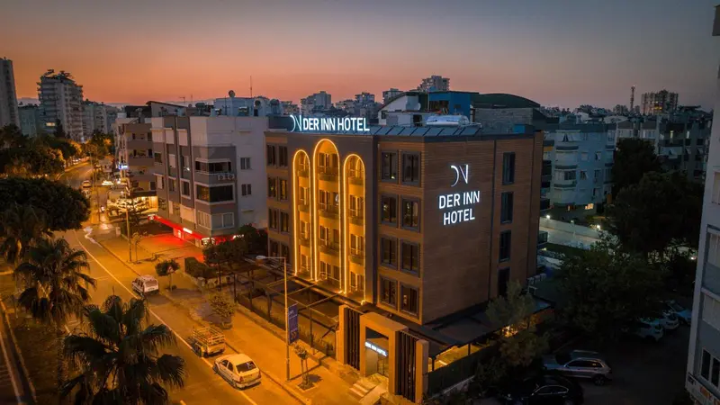 Vue aérienne d'un bâtiment moderne illuminé au crépuscule, avec des palmiers et des rues en contrebas.