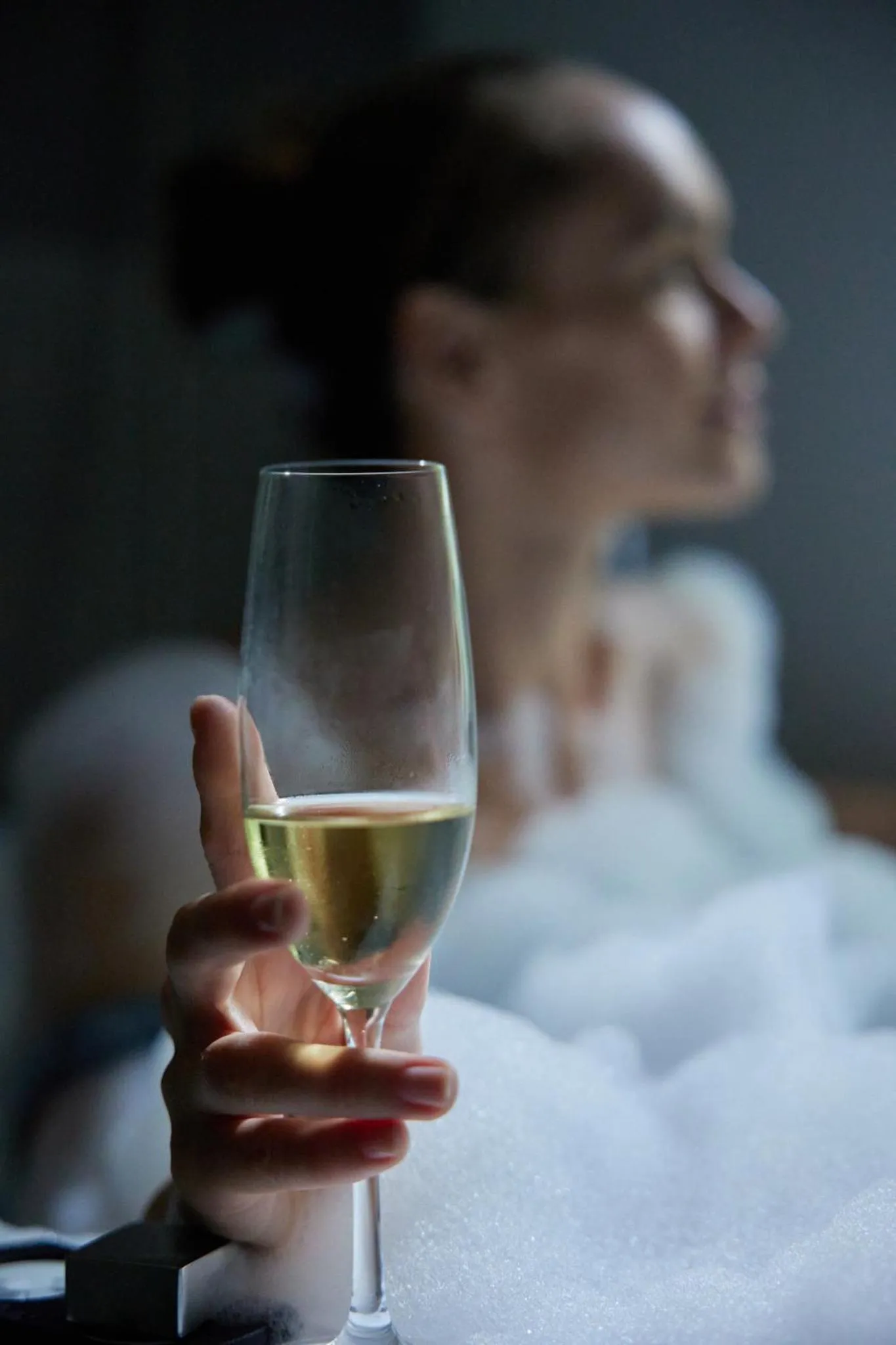 Une main tenant un verre de vin blanc au premier plan, une personne floue en arrière-plan.