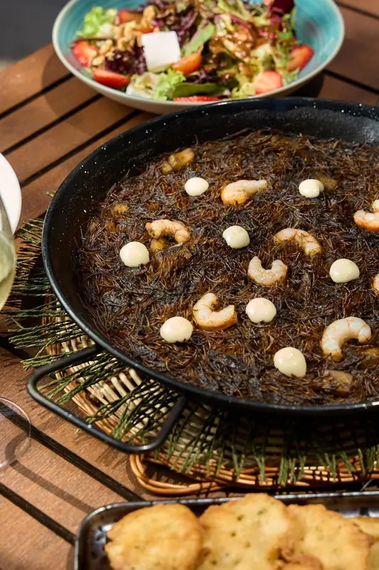 Une grande paella noire aux fruits de mer est présentée sur une table en extérieur.