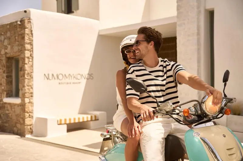 Un couple souriant est assis sur un scooter devant l'entrée d'un bâtiment blanc.