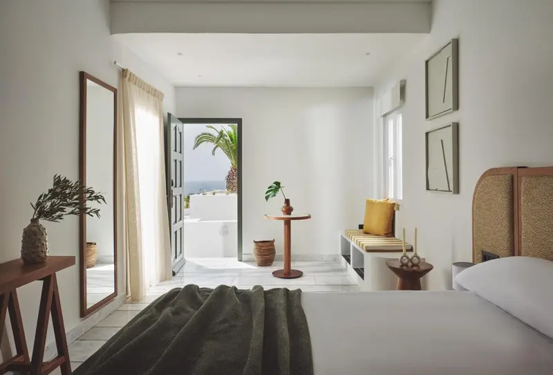 Chambre lumineuse avec un lit blanc et une vue sur la mer par une porte ouverte.