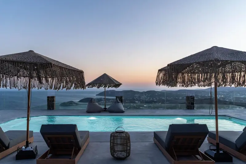 Terrasse avec transats et parasols face à une piscine et un paysage montagneux au coucher du soleil.