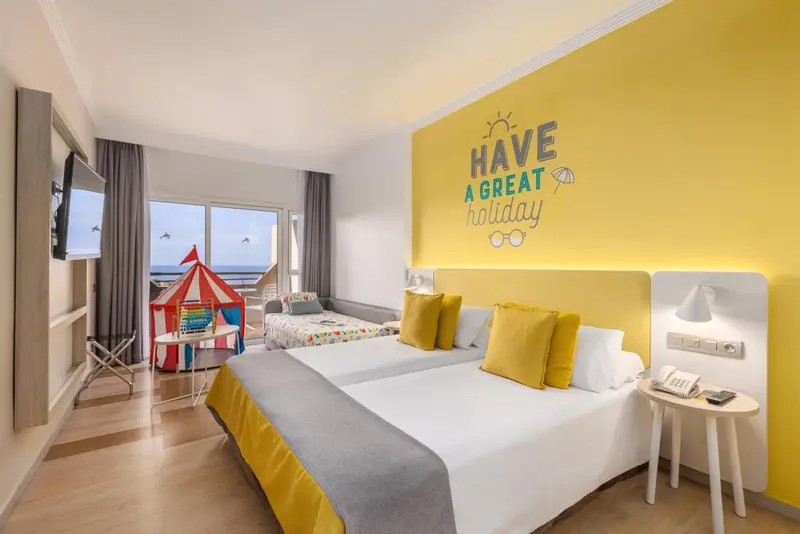 Chambre lumineuse avec un lit double et un lit simple, décorée de coussins jaunes et d'un mur jaune vif.