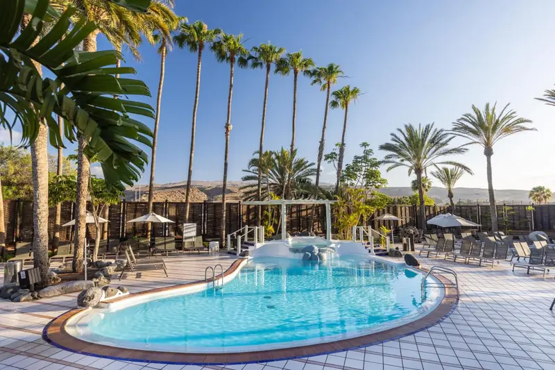 Une piscine rectangulaire bordée de chaises longues et de palmiers sous un ciel bleu.