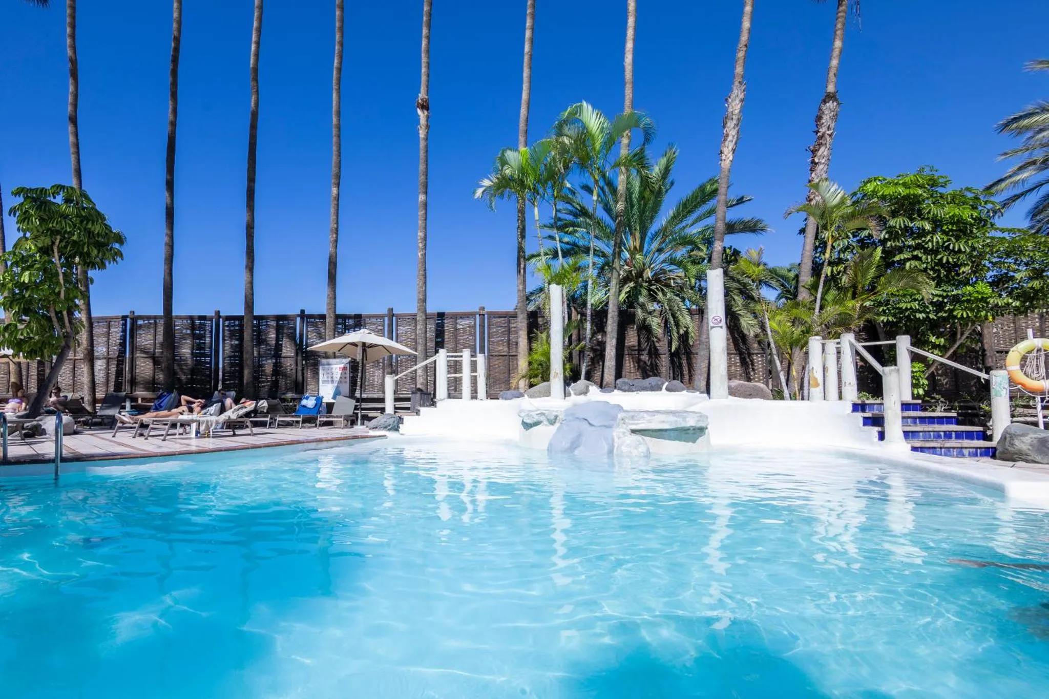 Une piscine turquoise bordée de transats et de palmiers sous un ciel bleu.
