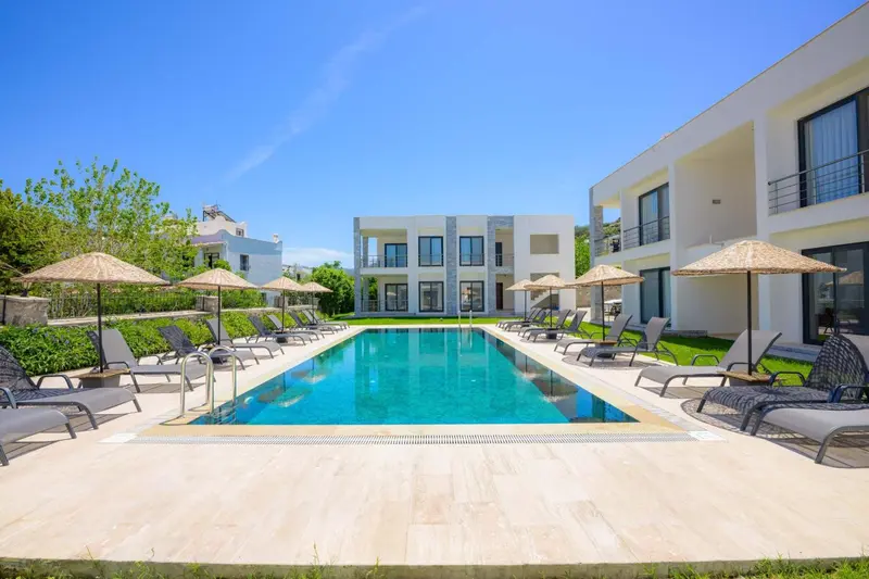 Une piscine rectangulaire turquoise est bordée de transats et de parasols devant un bâtiment blanc.