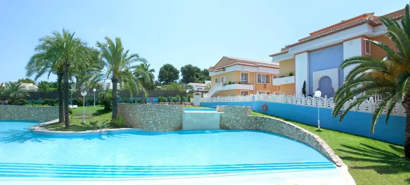 Une piscine turquoise avec une cascade artificielle et une pelouse verdoyante devant des bâtiments colorés.