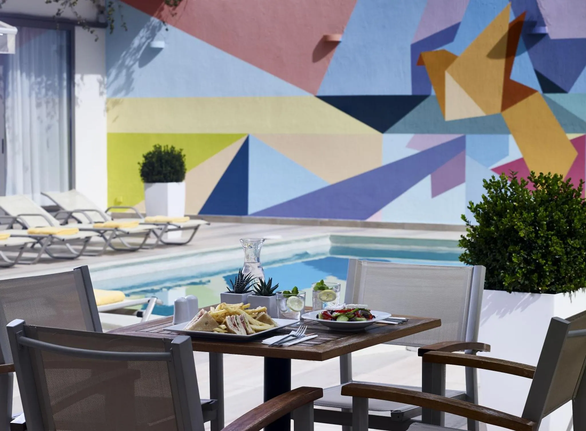 Une table est dressée avec des plats et des boissons près d'une piscine et d'une décoration murale colorée.