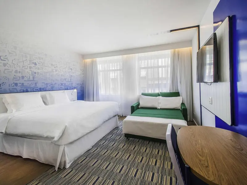 Chambre lumineuse avec un lit blanc et un canapé vert près d'une fenêtre.