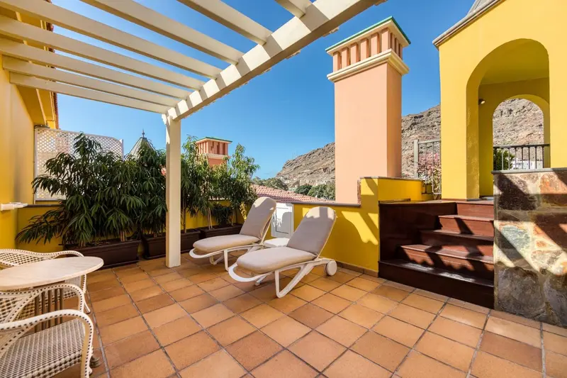 Terrasse ensoleillée avec transats et table basse, vue sur un paysage montagneux.