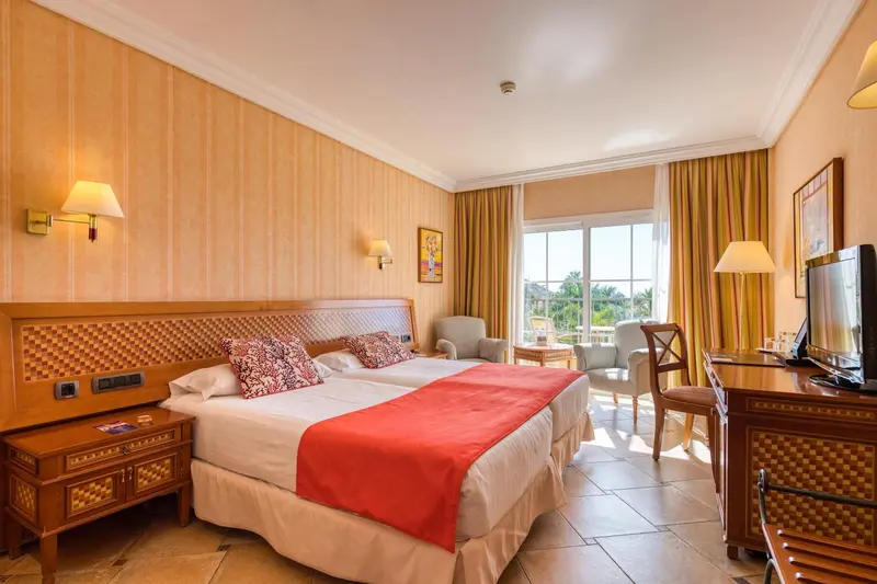 Chambre lumineuse avec un grand lit et un coin salon près de la fenêtre.