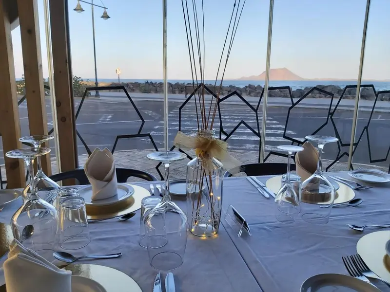 Une table dressée avec des couverts et des verres, devant une grande baie vitrée donnant sur la mer.