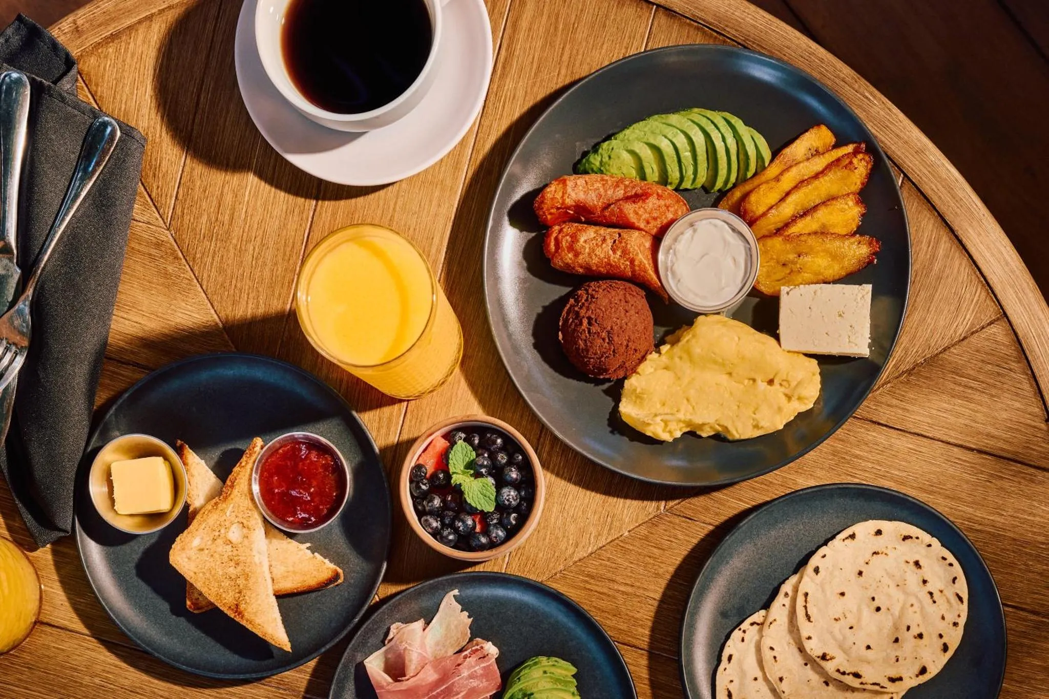 Une table ronde en bois présente un petit-déjeuner copieux avec des œufs, des saucisses, des fruits et des boissons.