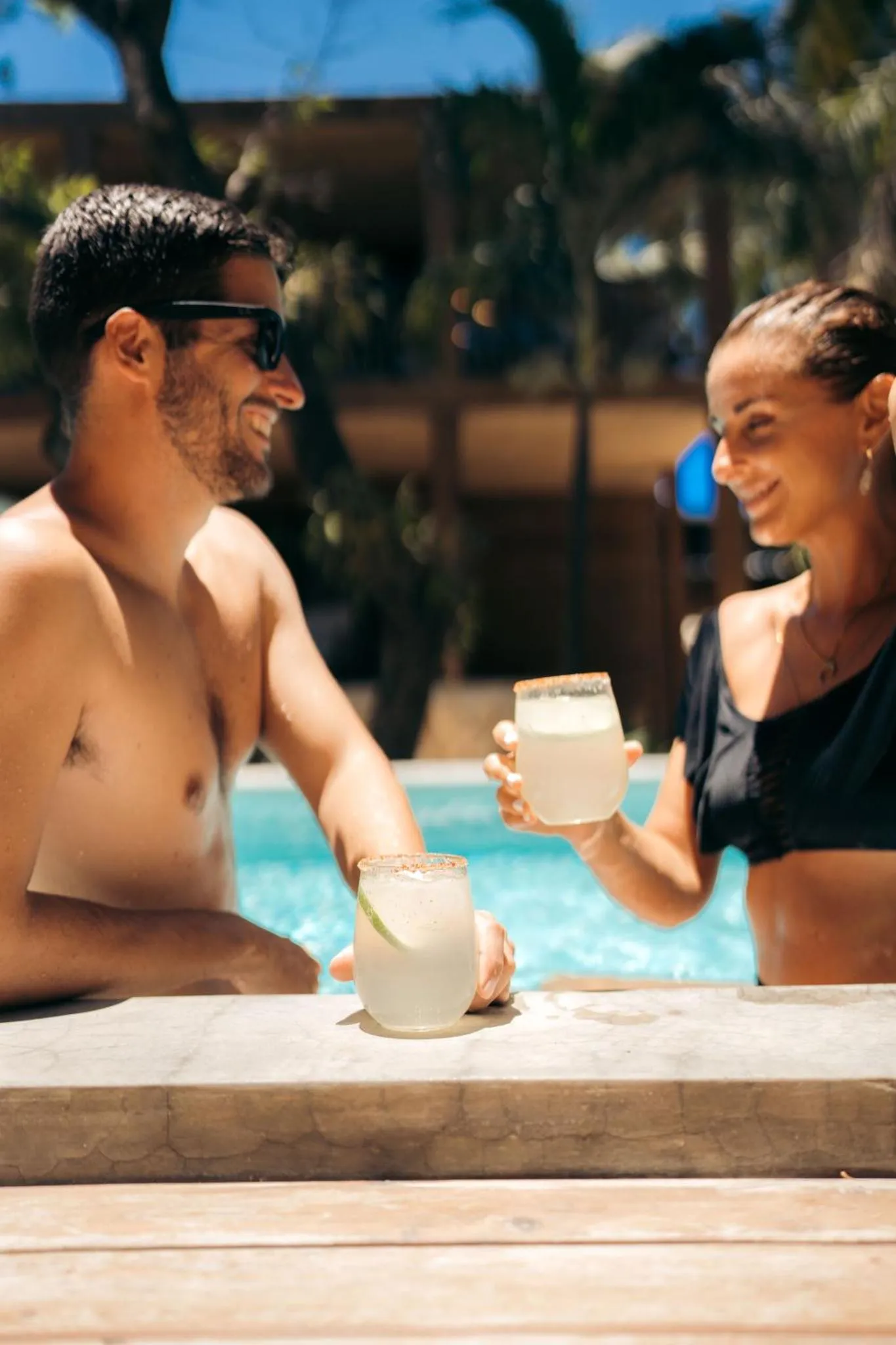 Un couple souriant trinque avec des cocktails au bord d'une piscine.