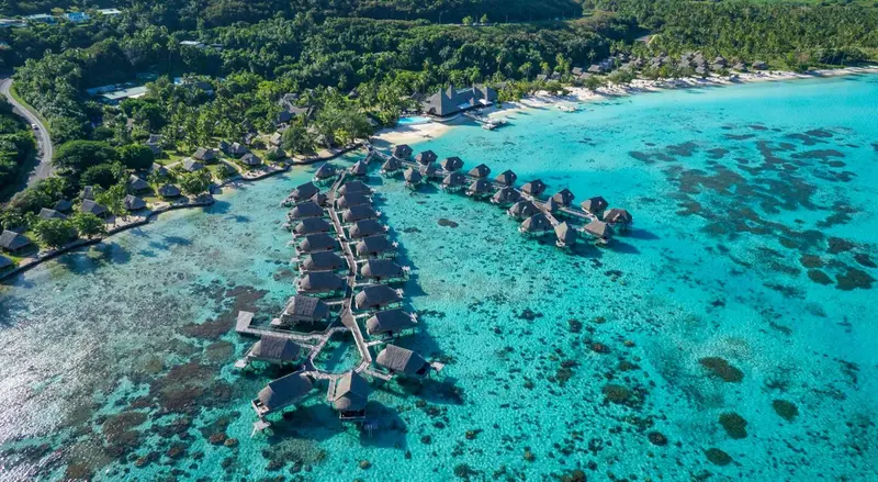 Vue aérienne d'un ensemble de bungalows sur pilotis au-dessus d'un lagon turquoise.