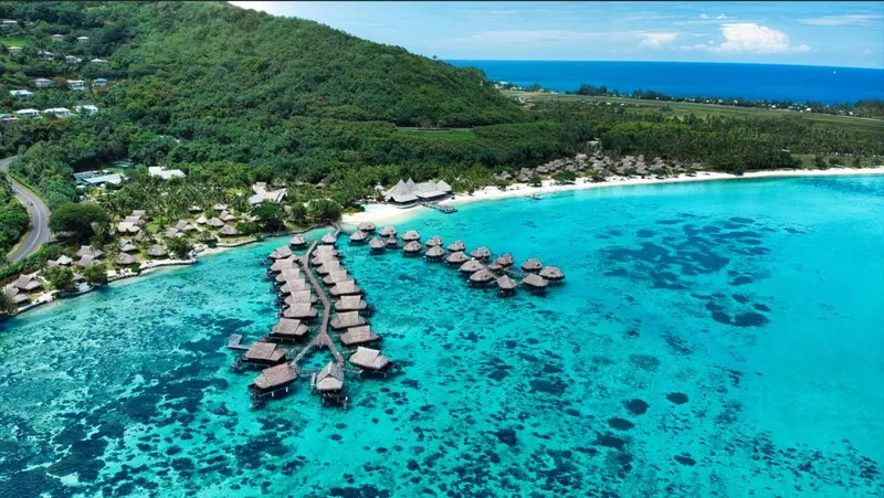 Vue aérienne d'une plage bordée de bungalows sur pilotis et d'une végétation luxuriante.
