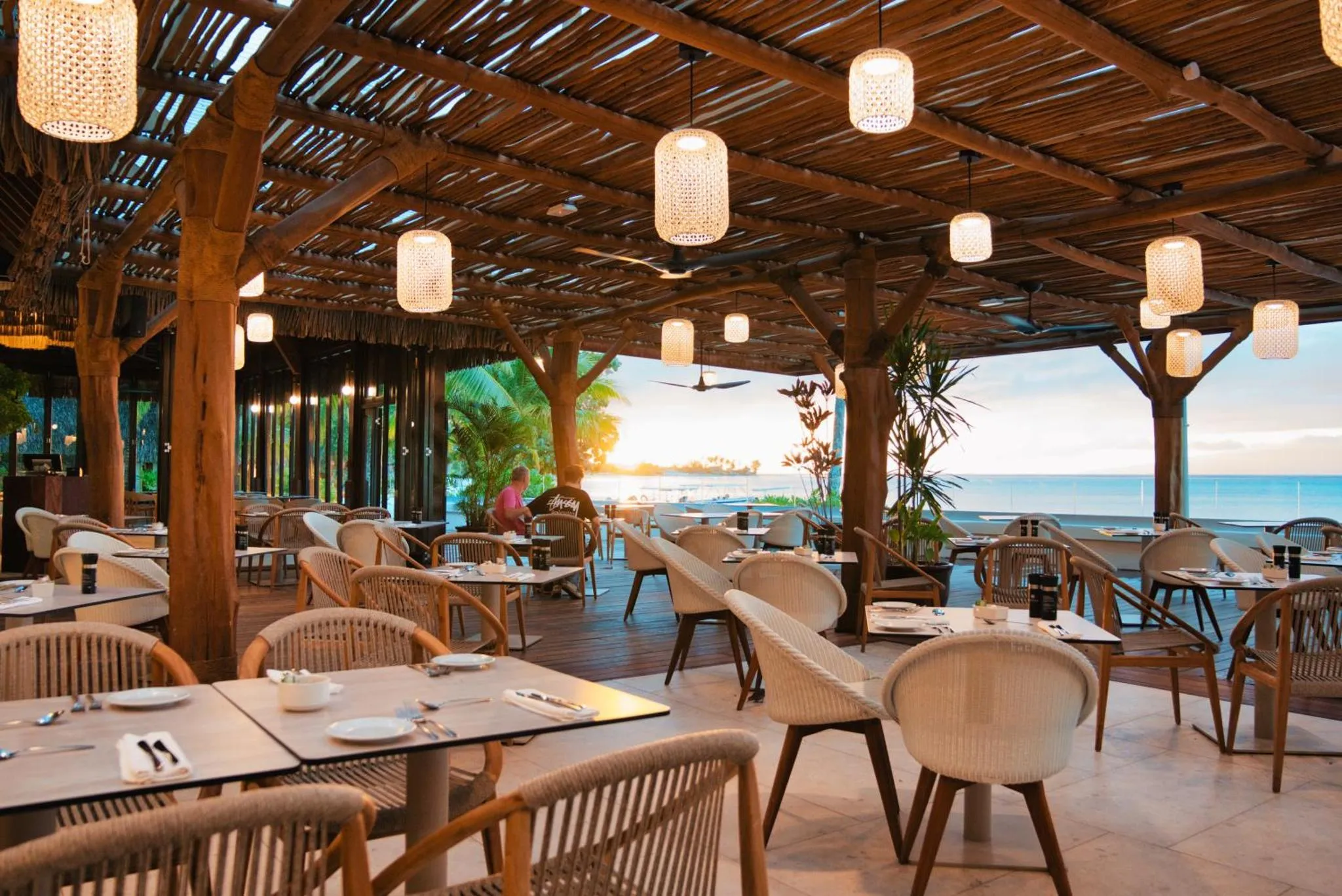 Terrasse ombragée avec tables et chaises face à la mer au coucher du soleil.
