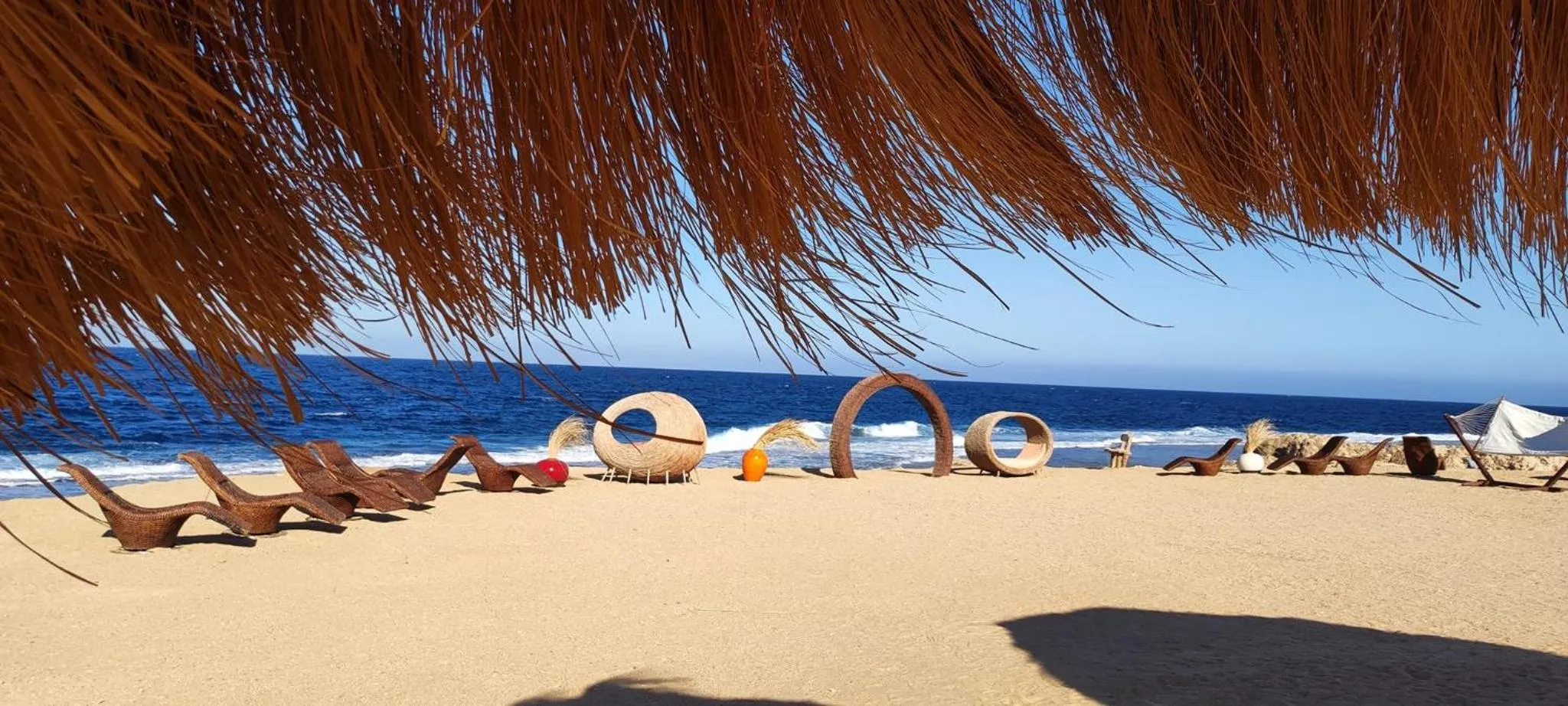 Transats et mobilier de plage sous un abri de palmiers face à la mer.