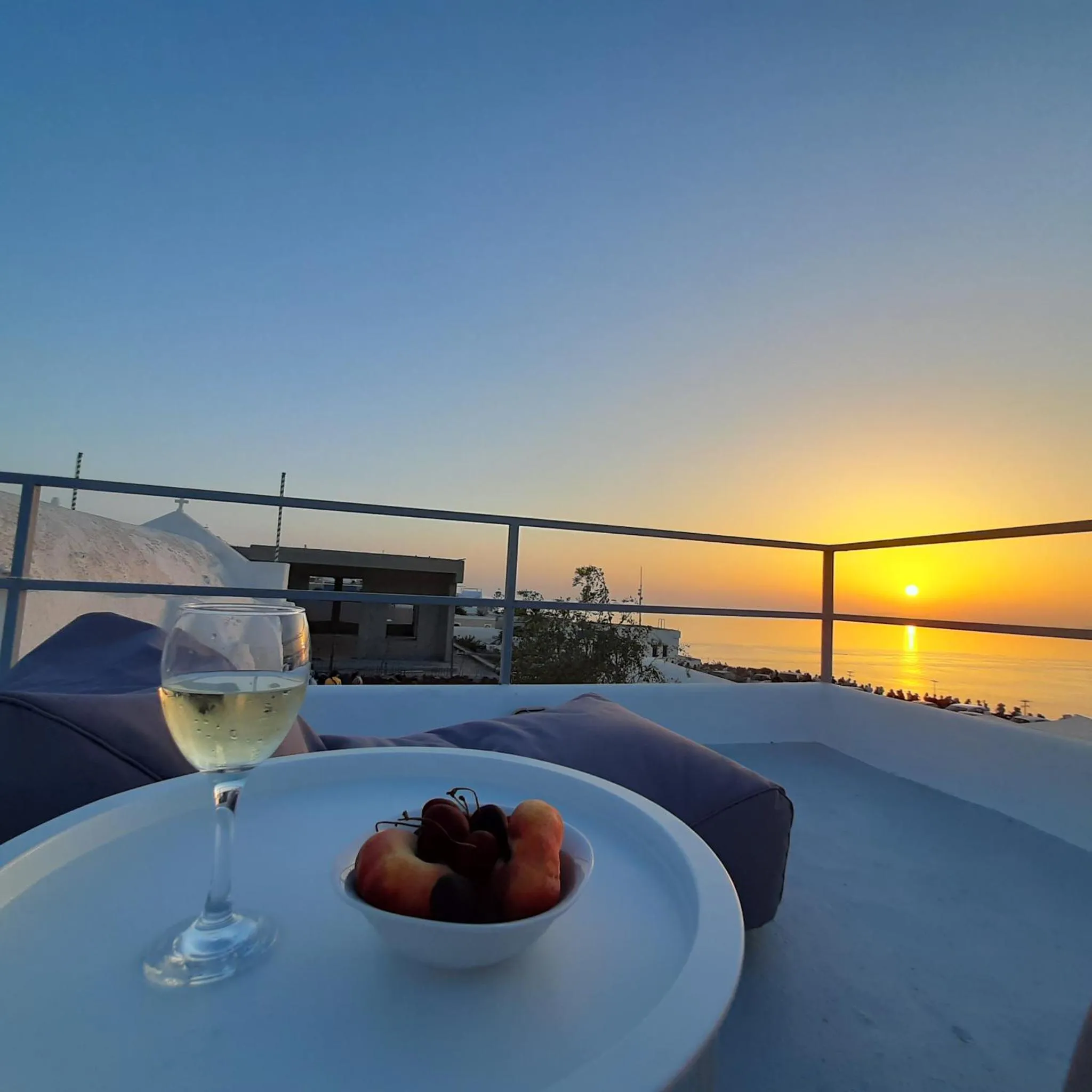 Une table basse avec des fruits et un verre de vin est posée sur une terrasse avec vue sur la mer au coucher du soleil.