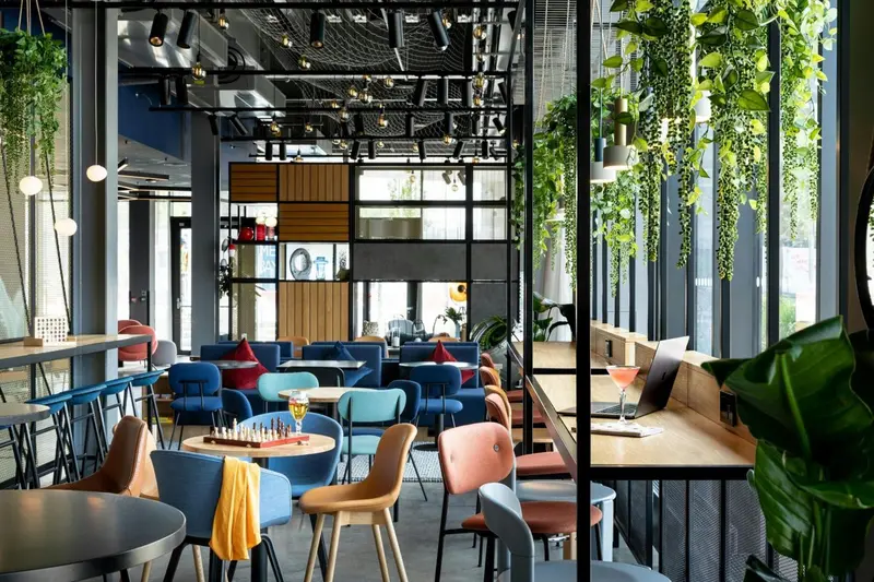 Une salle lumineuse avec des tables et des chaises colorées, des plantes suspendues et de grandes fenêtres.