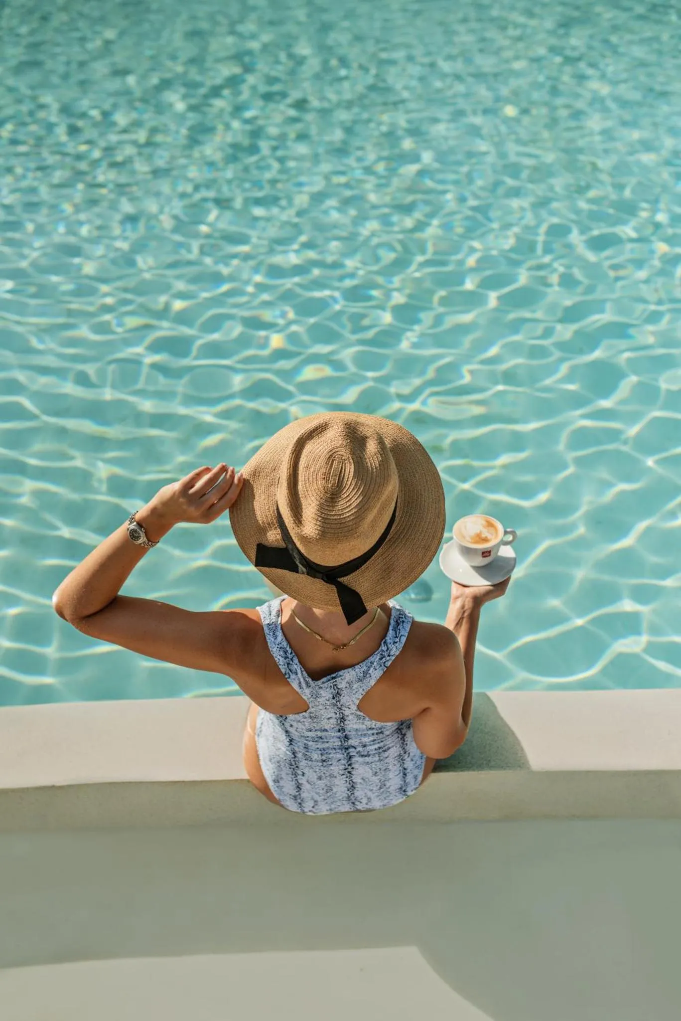 Une personne assise au bord d'une piscine, un chapeau de paille et une boisson à la main.