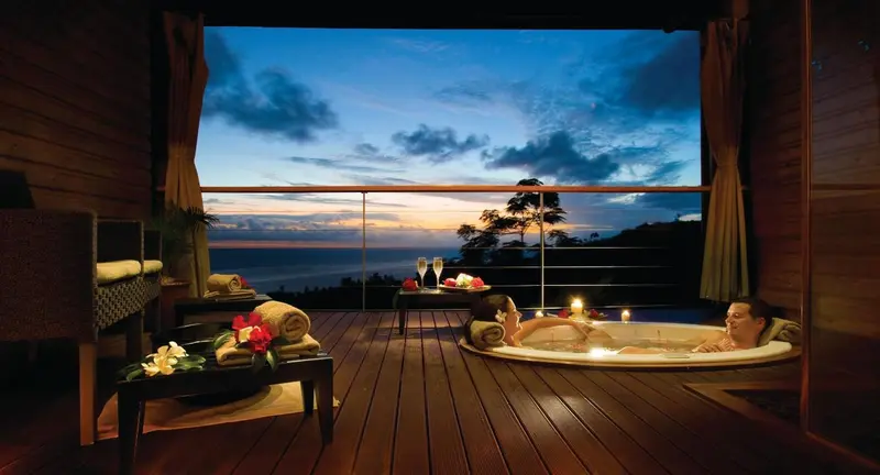 Un couple se détend dans un jacuzzi sur une terrasse avec vue sur la mer au coucher du soleil.