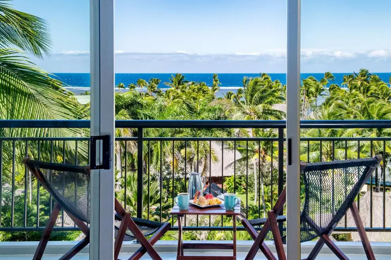 Deux fauteuils et une table basse avec des fruits et des boissons sur un balcon avec vue sur la mer et la végétation.