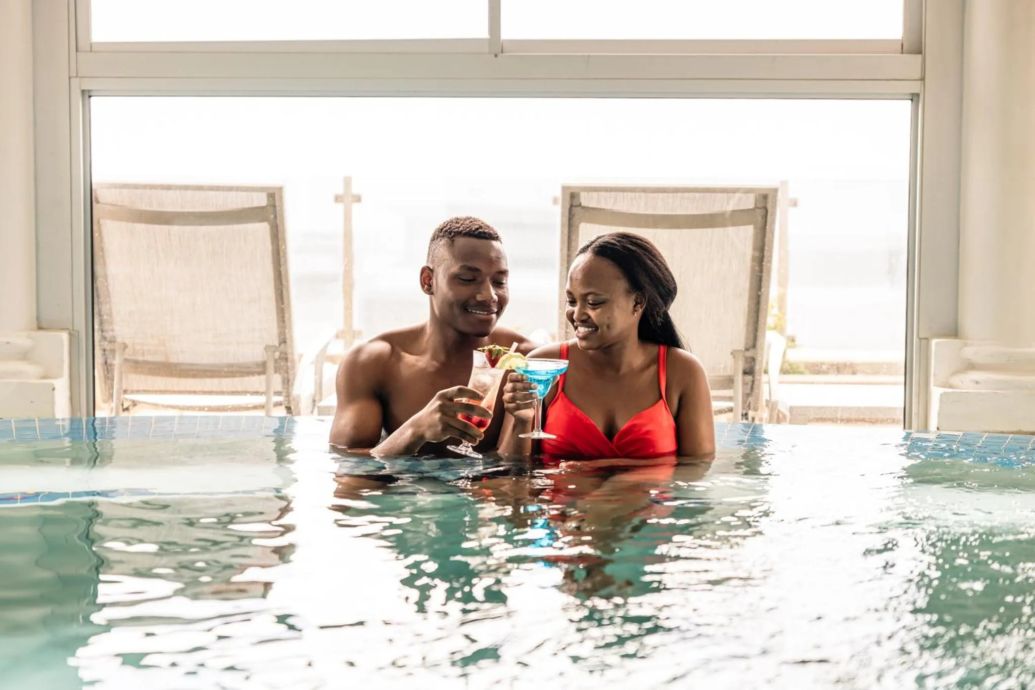 Un couple souriant profite de cocktails dans une piscine avec vue sur la mer.