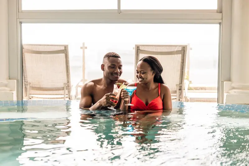 Un couple souriant profite de cocktails dans une piscine avec vue sur la mer.
