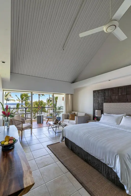 Une chambre lumineuse avec un lit fait et un coin salon donnant sur une vue tropicale.