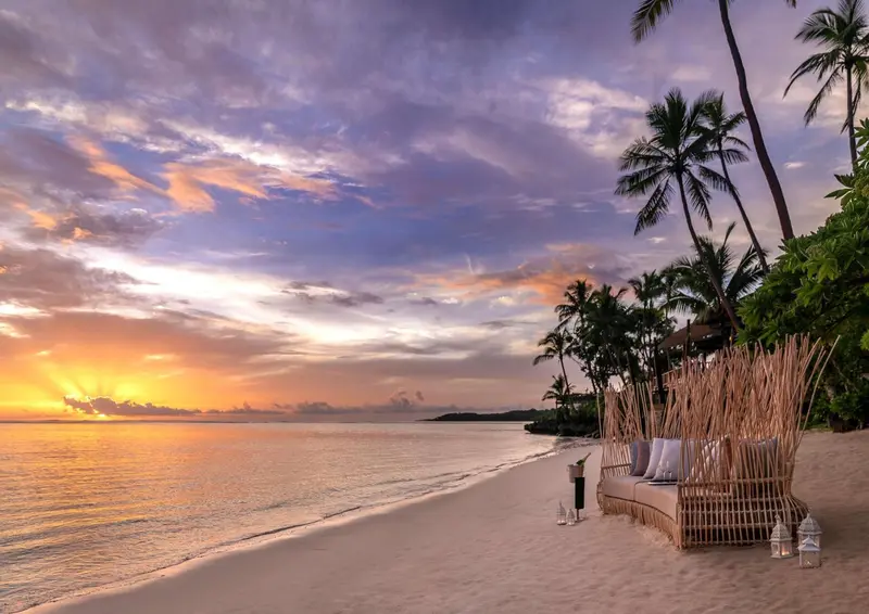 Coucher de soleil sur une plage de sable fin avec des palmiers et des chaises longues.