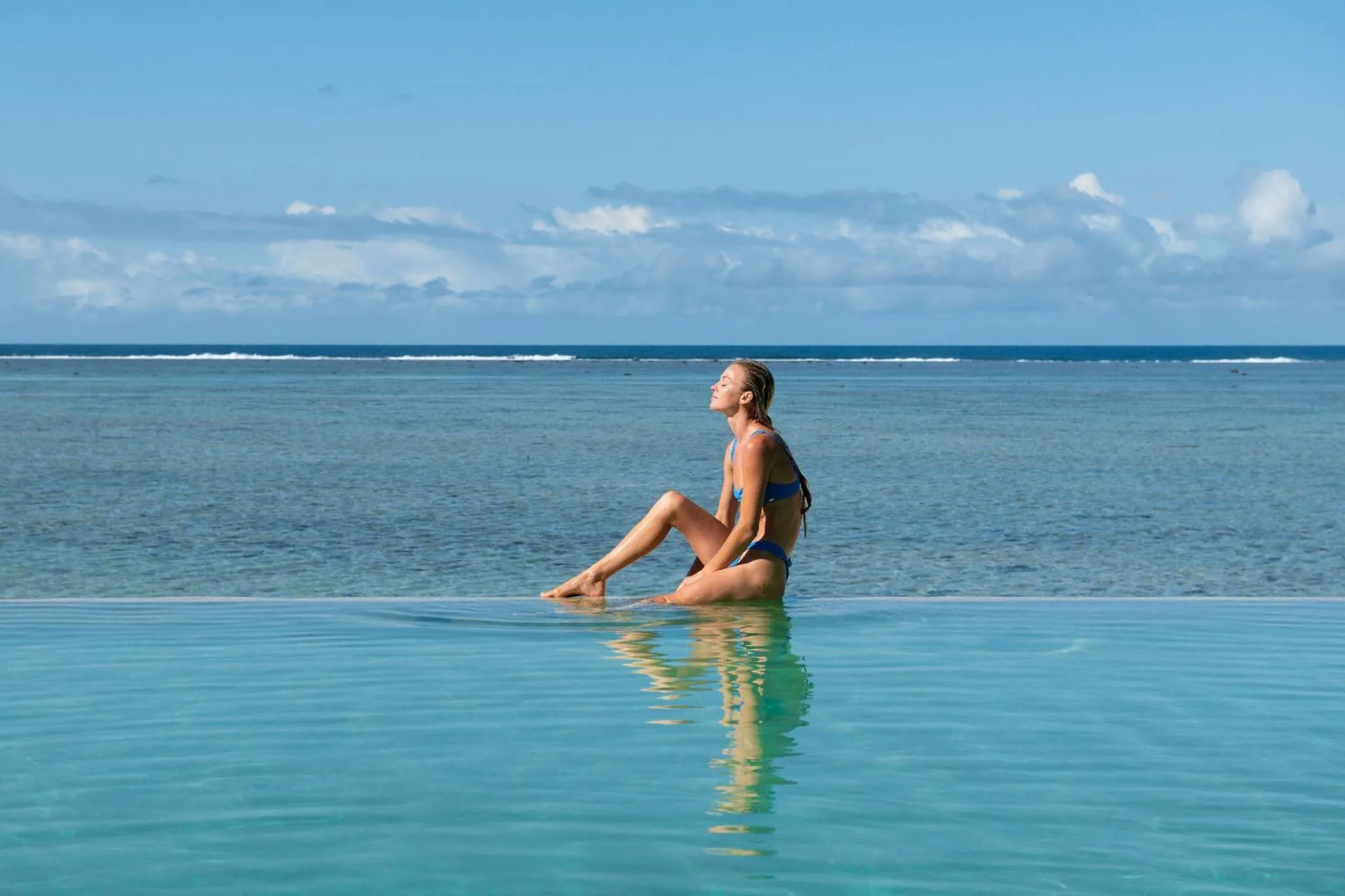 Une femme est assise au bord d'une piscine à débordement face à la mer.