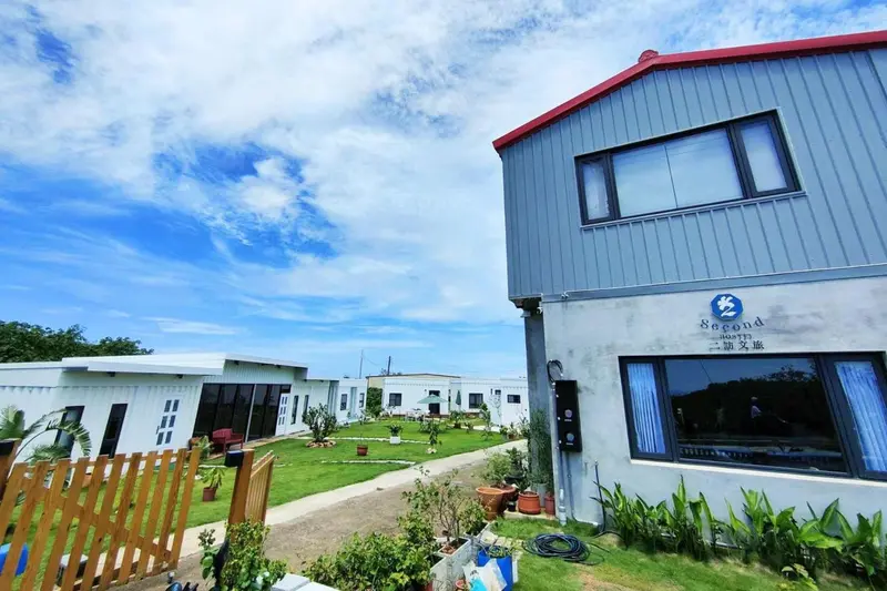 Bâtiment blanc à toit rouge avec pelouse et végétation environnante sous un ciel nuageux.