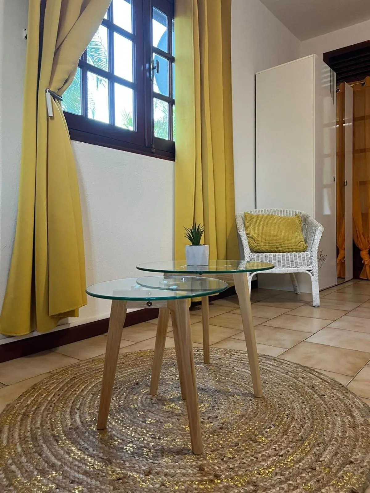 Une petite table ronde en verre et une chaise en osier se trouvent près d'une fenêtre avec des rideaux jaunes.