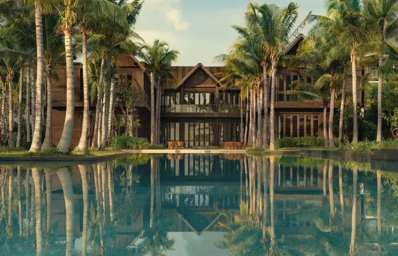 Bâtiment en bois sombre reflété dans une piscine bordée de palmiers.