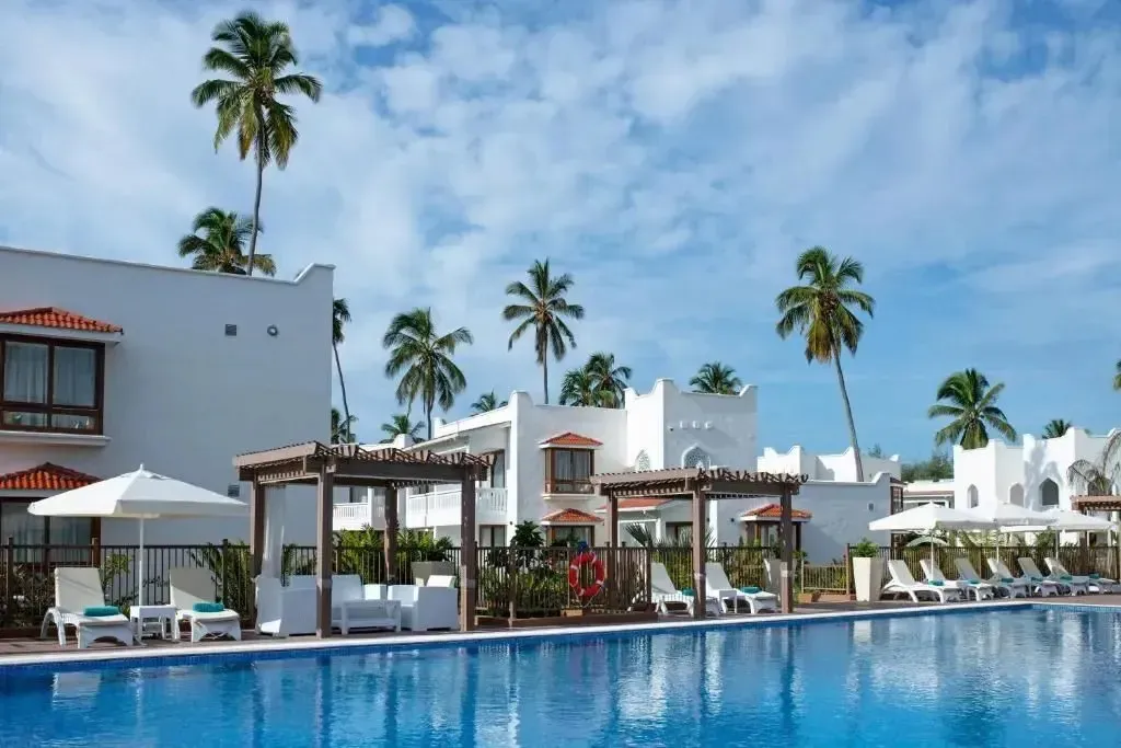 Une piscine turquoise bordée de transats et de parasols blancs, avec des bâtiments blancs et des palmiers en arrière-plan.
