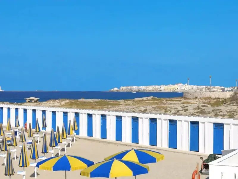Plage de sable avec parasols colorés et cabines de plage à rayures bleues et blanches.