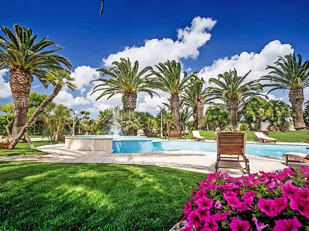 Une piscine rectangulaire avec une fontaine et des transats, entourée d'une pelouse verte et de palmiers.