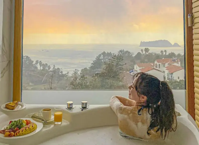 Une personne se détend dans une baignoire face à une vue sur la mer et un paysage verdoyant.