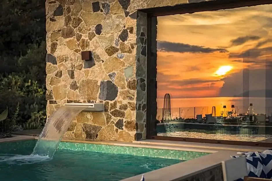 Une piscine avec cascade à débordement offre une vue sur un coucher de soleil orangé en mer.