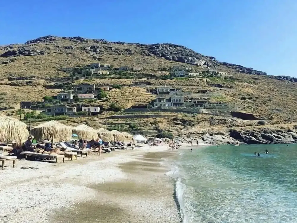 Une plage de sable fin bordée de parasols et de transats, avec des bâtiments sur une colline en arrière-plan.