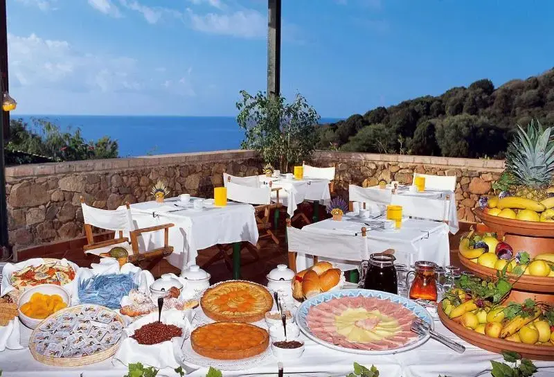 Une terrasse ombragée offre une vue sur la mer et la végétation environnante, avec une table garnie de mets variés.