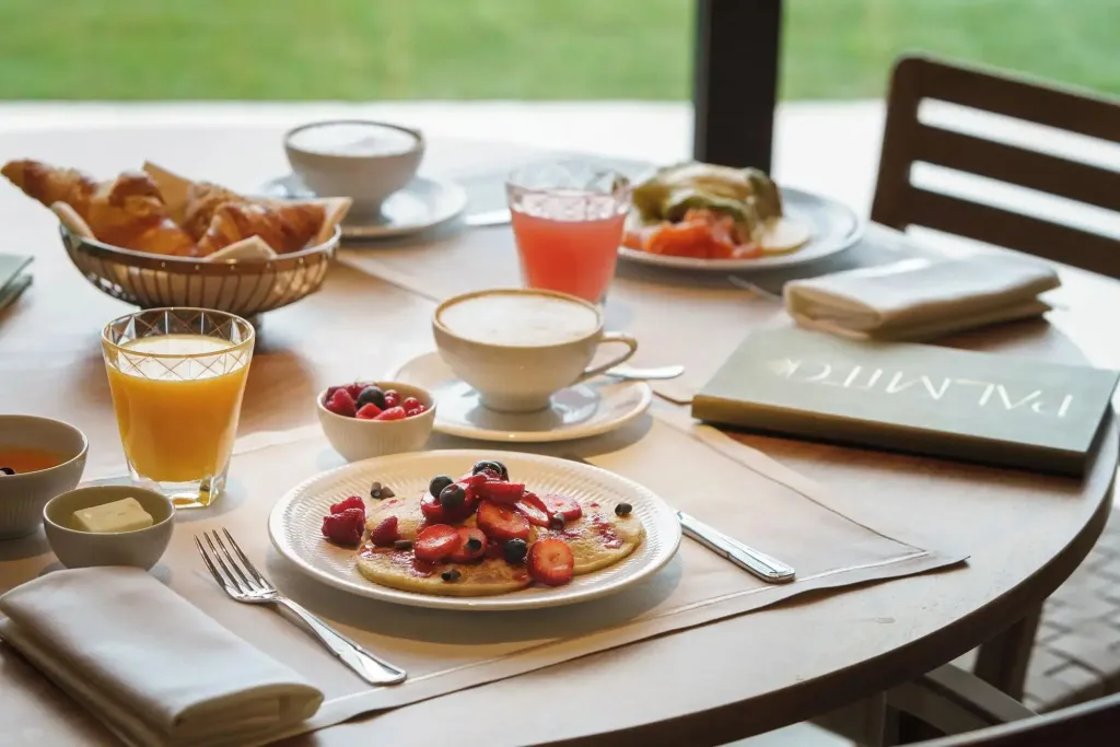 Une table ronde est dressée avec un petit-déjeuner composé de viennoiseries, de fruits et de boissons.