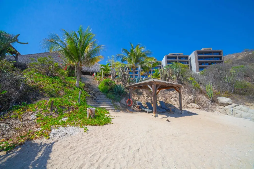 Une plage de sable fin mène à un escalier bordé de végétation tropicale et à un abri en bois.