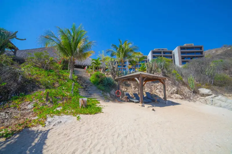 Une plage de sable fin mène à un escalier bordé de végétation tropicale et à un abri en bois.