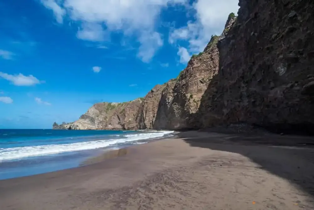 Une plage de sable sombre bordée par des falaises rocheuses sous un ciel bleu.