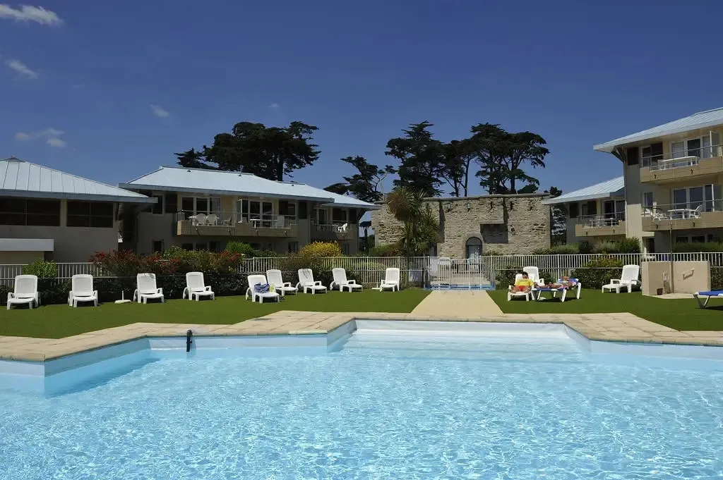Une piscine turquoise au premier plan, avec des transats alignés sur une pelouse verdoyante et des bâtiments en arrière-plan.