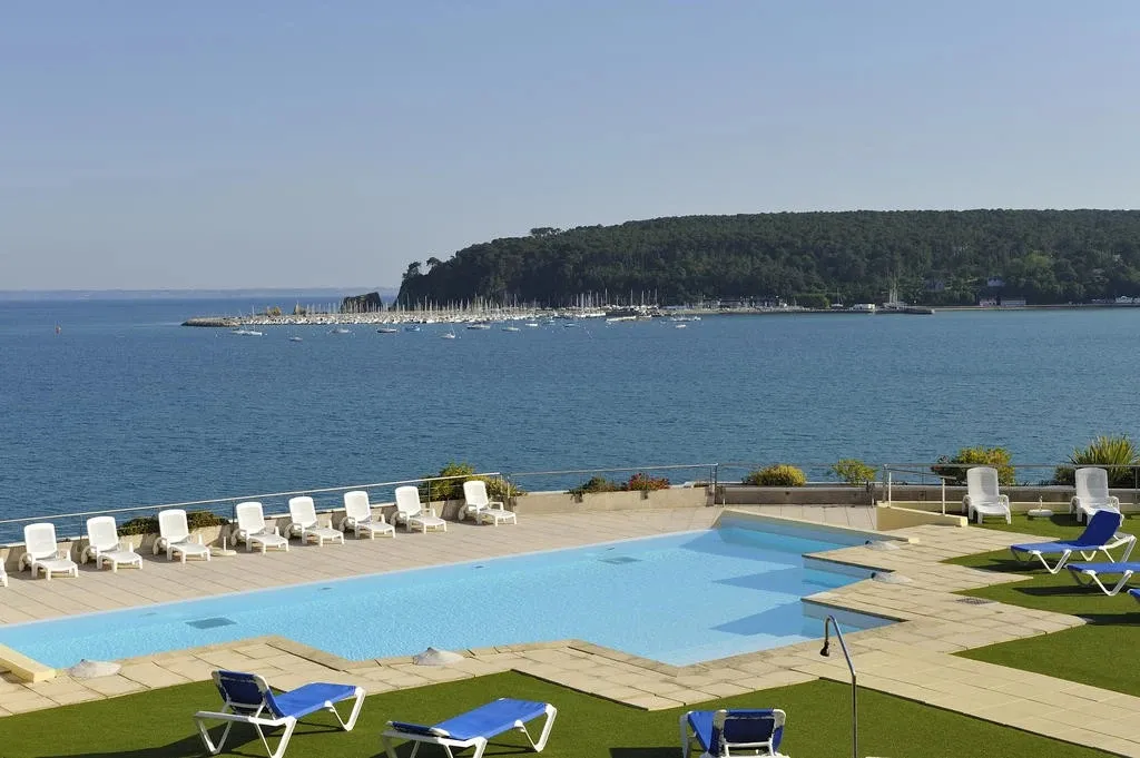 Une piscine avec des transats offre une vue sur la mer et un port de plaisance.